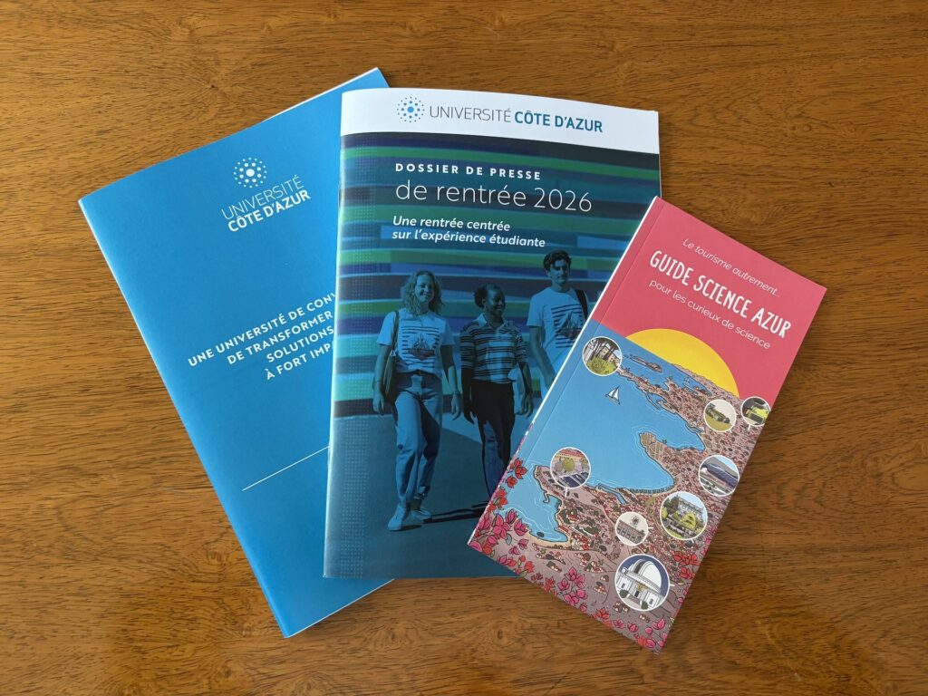 Three brochures from Côte d'Azur University placed on a wooden table: an institutional blue leaflet, the press kit for the 2026 academic year with three students on the cover, and the illustrated "Guide Science Azur".